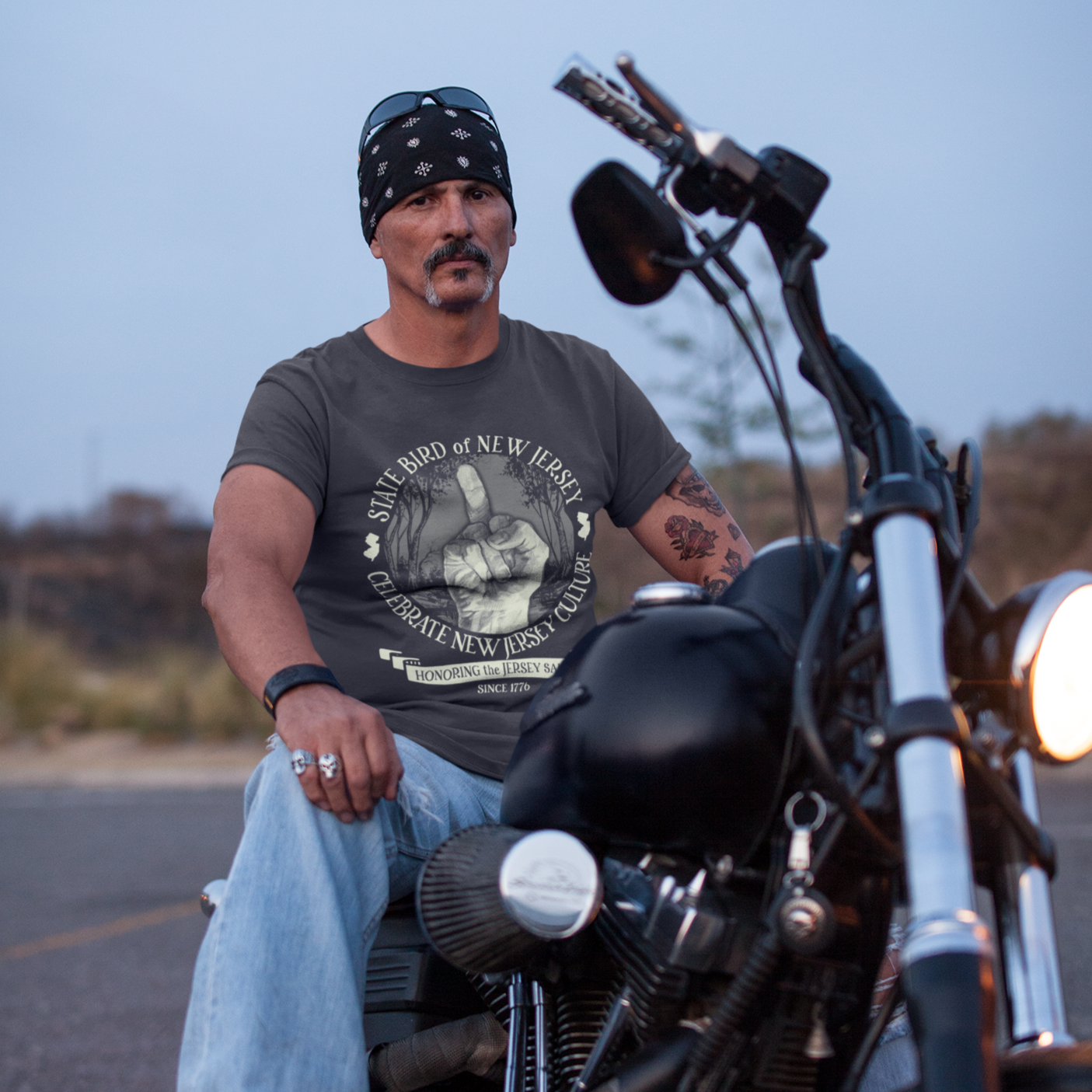 Man sitting on a motorcycle wearing a bandana and casual clothing, with a scenic background.